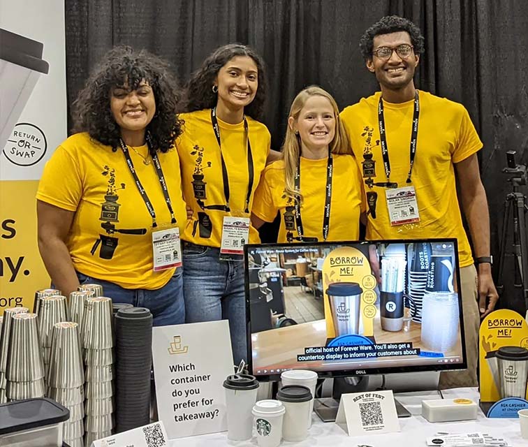 The Forever Ware team stands by their booth at Coffee Fest in Chicago wearing yellow shirts behind a table filled with reusable takeaway containers.