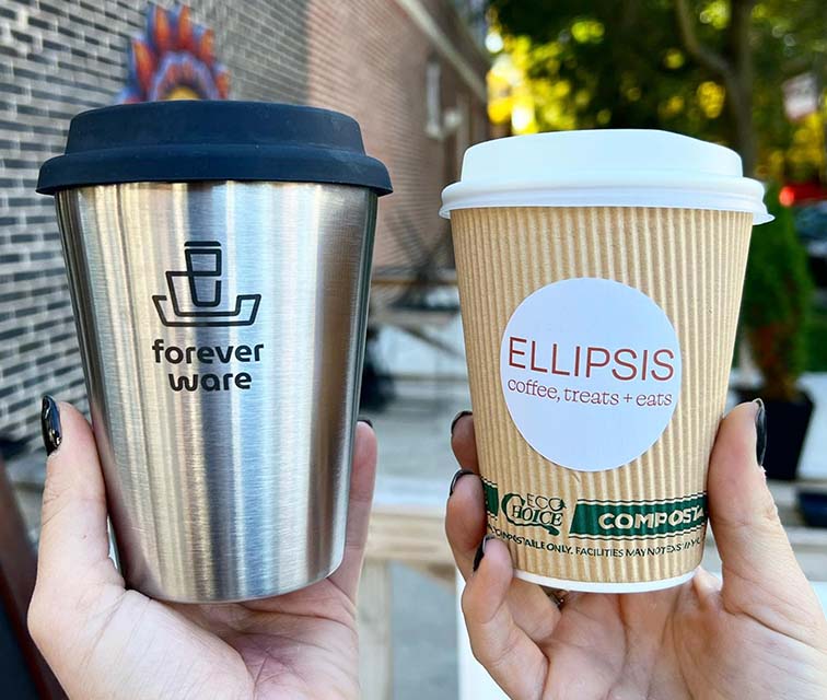 A Forever Ware reusable stainless steel mug with a black silicone lid held up beside a compostable paper cup branded with Ellipsis Coffee, treats + eats.